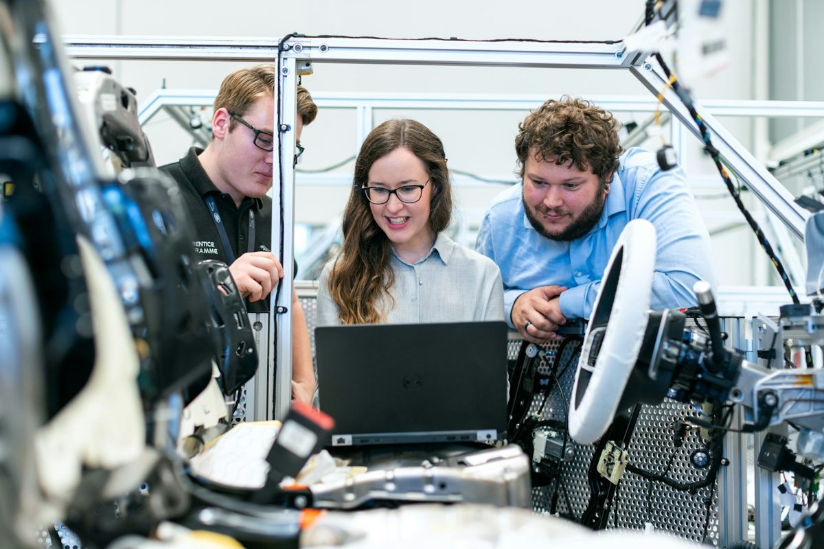 Three engineering students reviewing scenario data on a laptop in a vehicle assembly workspace